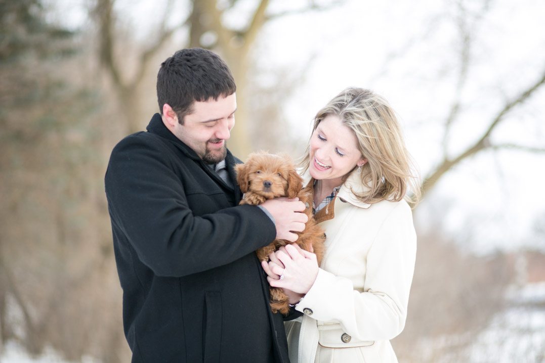 couple smiling at their puppy in their winter engagement photos in Chippewa Falls