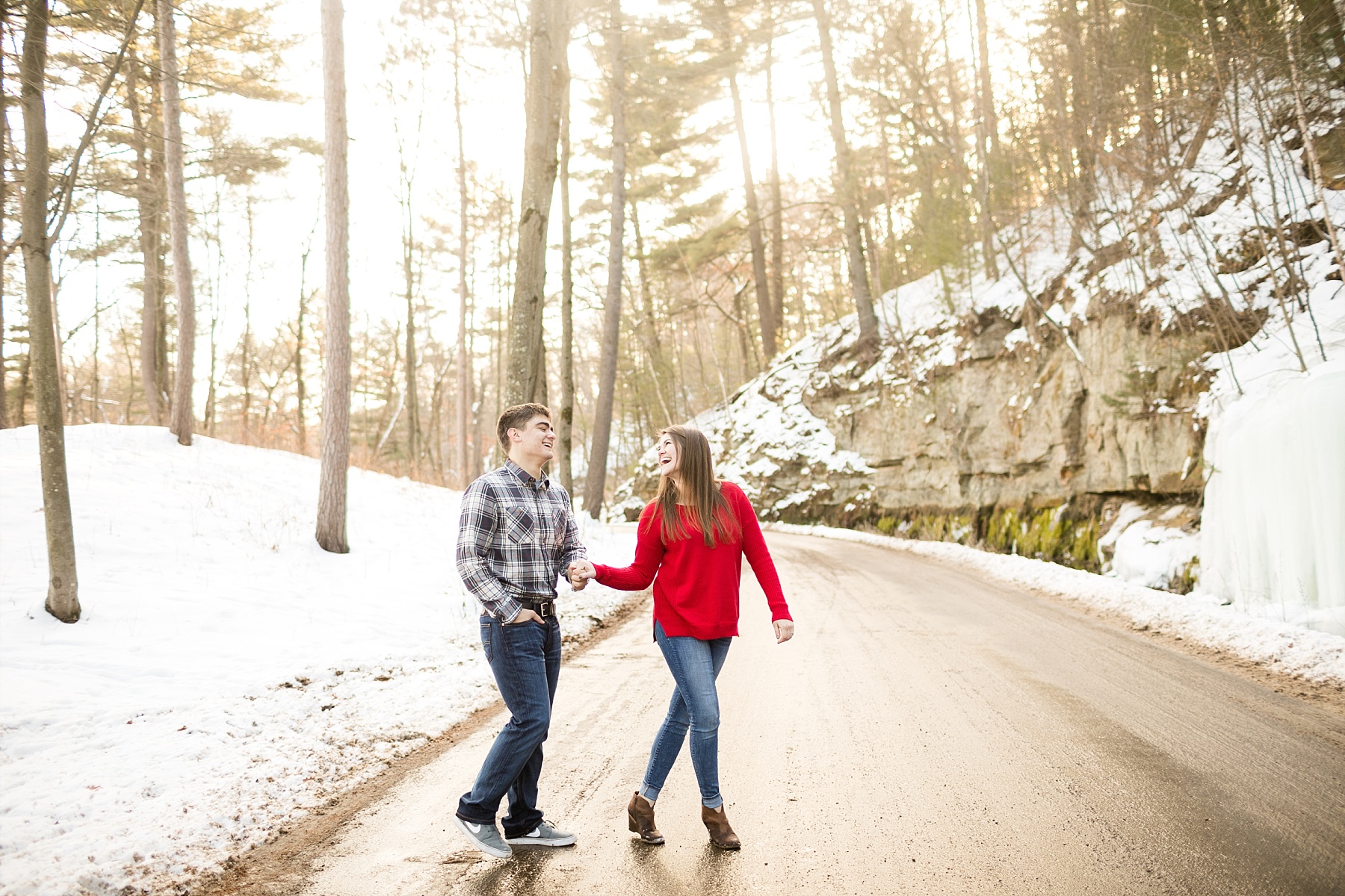 girl in a red sweater looking behind to her fiance in their winter engagement photos in Chippewa Falls