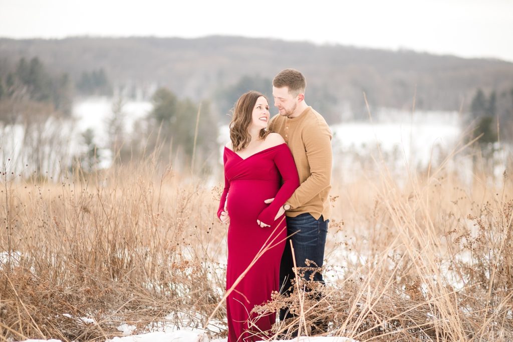 couple in a field with tall grass and snow smiling at each other for their maternity photos