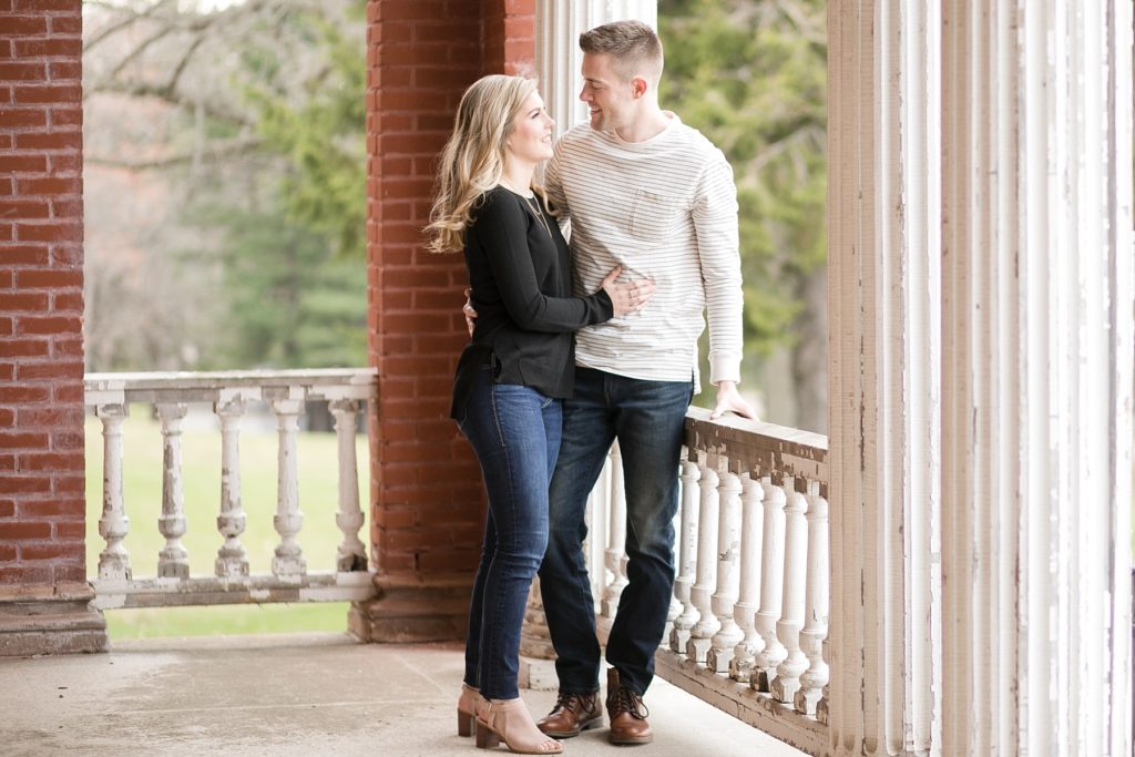 Rachel and AJ standing on the covered porch of an abandoned building at the Northern Colony in Chippewa Falls, WI for their engagement session