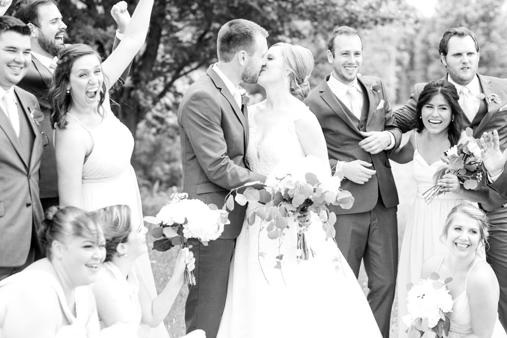 black and white photo of bride and groom kissing with bridal party cheering at wedding atThe Florian Gardens in Eau Claire