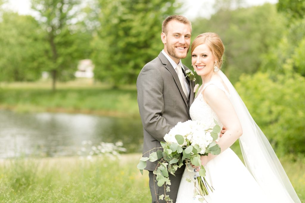 bride and groom smiling at the camera infront of a lake at wedding atThe Florian Gardens in Eau Claire