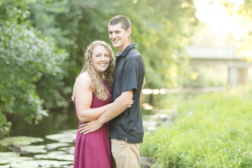 couple standing on rocks in a creek with the sun and a bridge behind them in Bloomer WI for their engagement photos