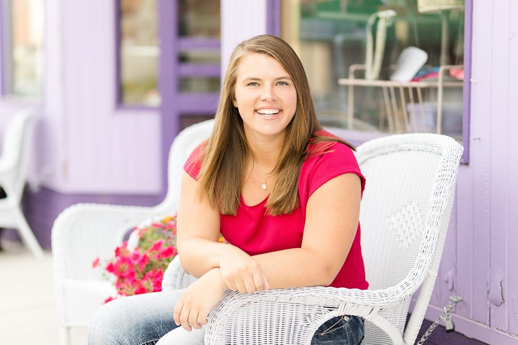 girl in hot pink shirt by a purple building sitting in a wicker chair for her senior photos in Chippewa Falls, WI