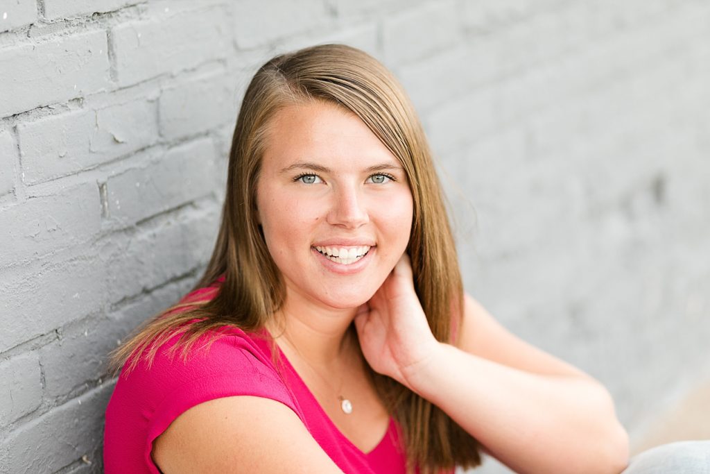 girl leaning on grey brick wall smiling at camera in a hot pink shirt for her senior photos in Chippewa Falls