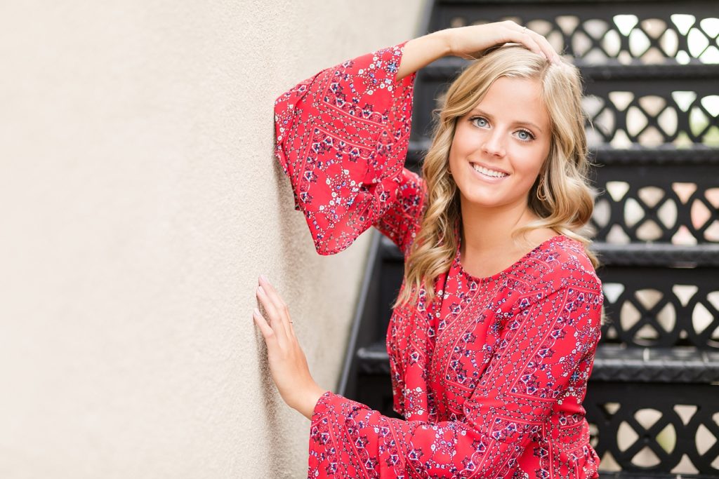 girl in boho red top sitting on a staircase with her hand over her head smiling for her Chippewa Falls senior photos