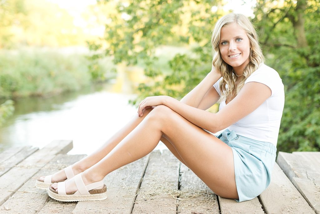 girl on an old farm bridge over a creek sitting smiling in blue shorts and a white tee in Bloomer WI for senior photos