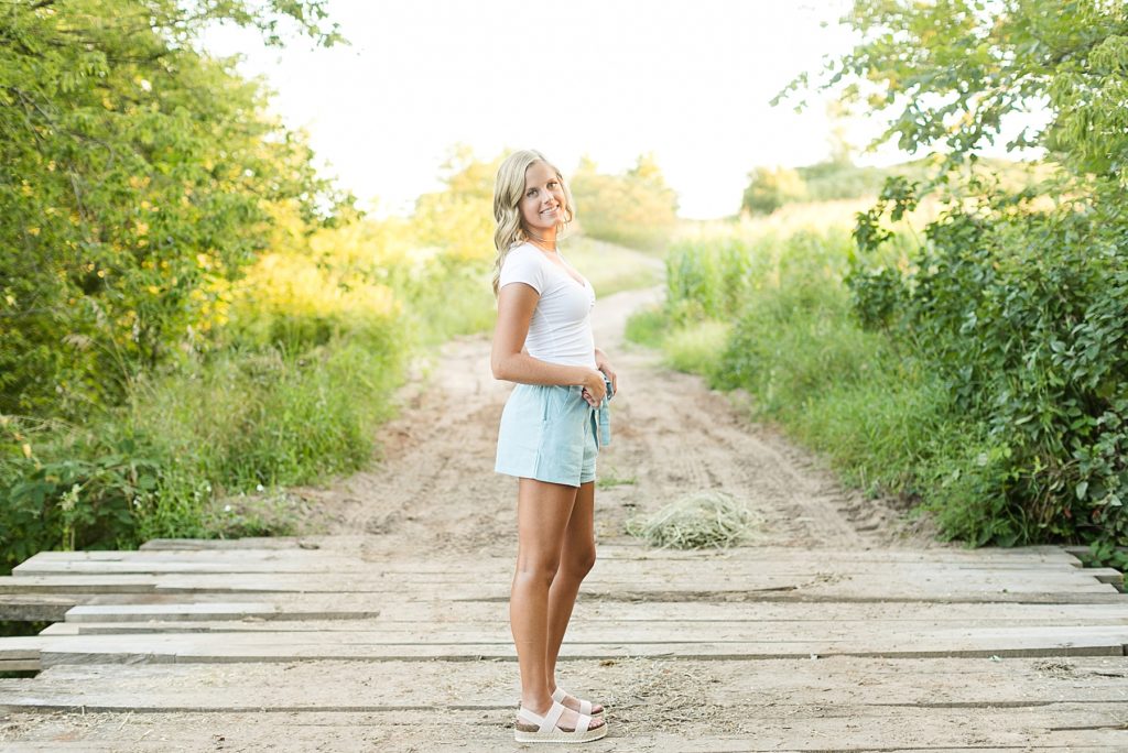 girl in a white shirt and blue shorts looking back standing on a bridge at their farm in Wisconsin for her senior photos