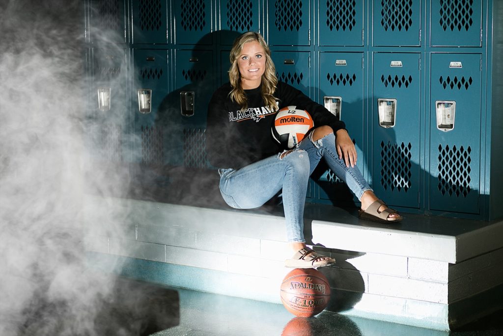 girl sitting in a locker room with her foot on a basketball and holding a volleyball and smoke coming over the left side of the photo in Bloomer WI