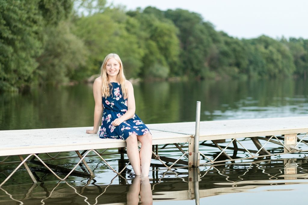girl sitting on a dock in a blue floral dress by Half Moon Lake in Eau Claire for her senior photos