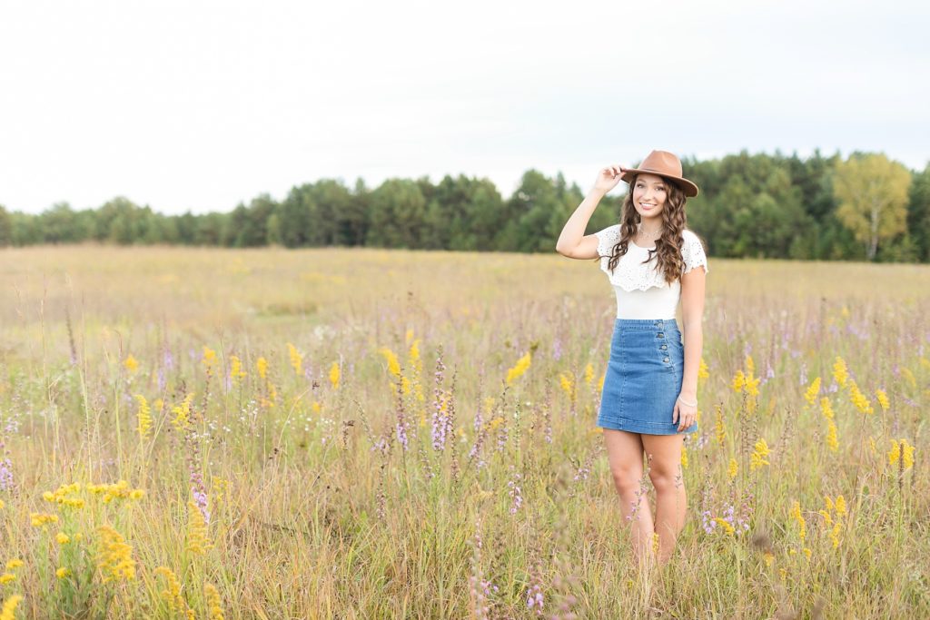 girl in a field of flowers in a denim skirt and white top with a brown hat smiling at Lake Wissota State Park for her senior photos