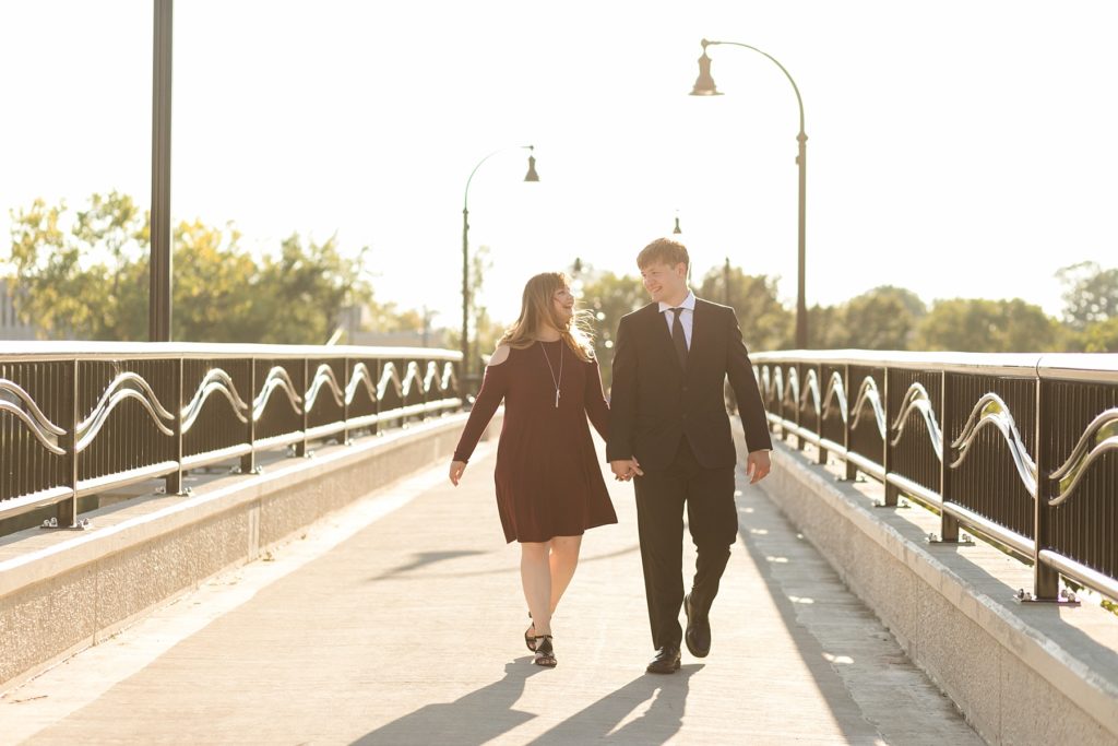 couple with the wind blowing in their hair walking across a footbridge in Eau Claire for their engagement pictures