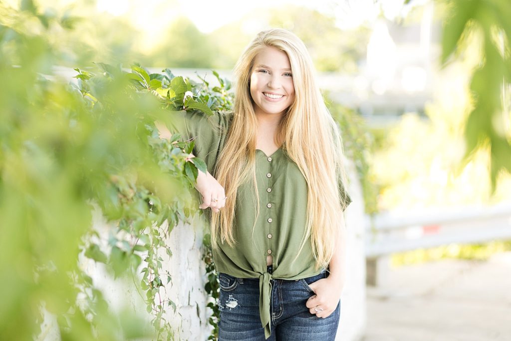 Rice Lake senior leans against a vine wall smiling in a green shirt and jeans.