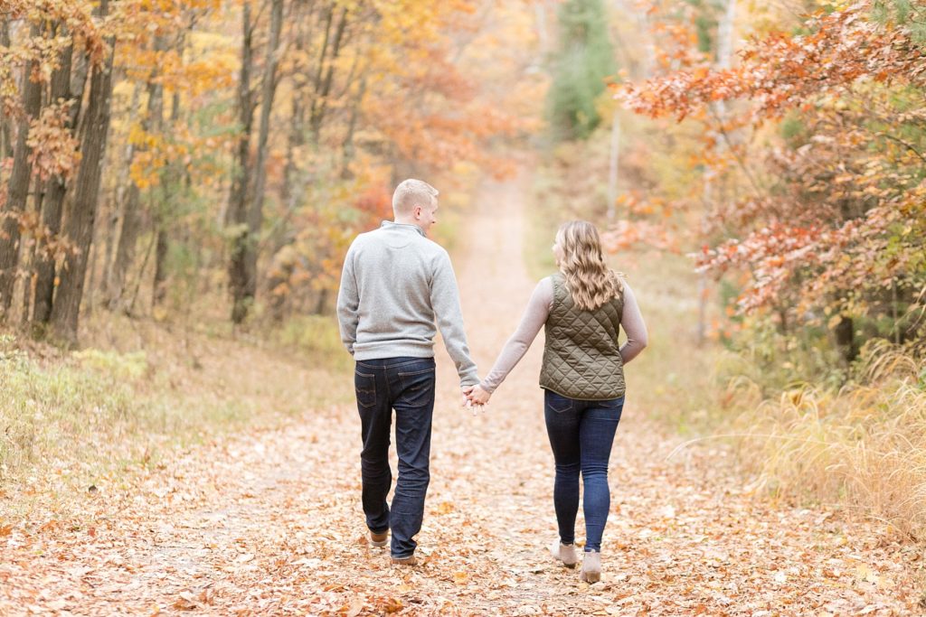 couple walking down a dirt road covered with fall leaves for their woodsy Wisconsin engagement session