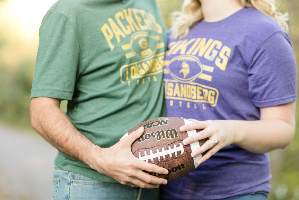 couple with Packers and Vikings shirts hold football with engagement ring showing