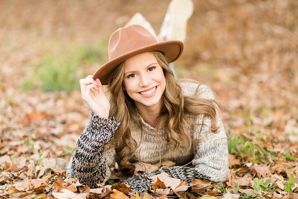 girl laying in brown leaves with a brown hat on for her fall Eau Claire senior photos at River Prairie