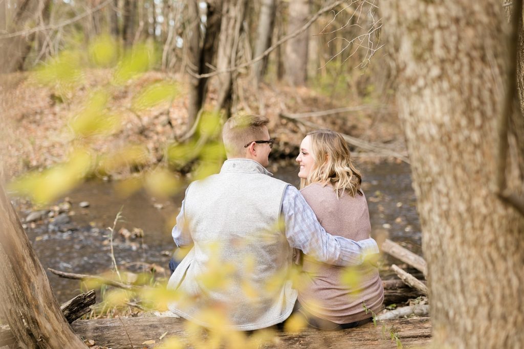 couple smiling at each other sitting on a log for their late fall Irvine Park engagement photos in Chippewa Falls