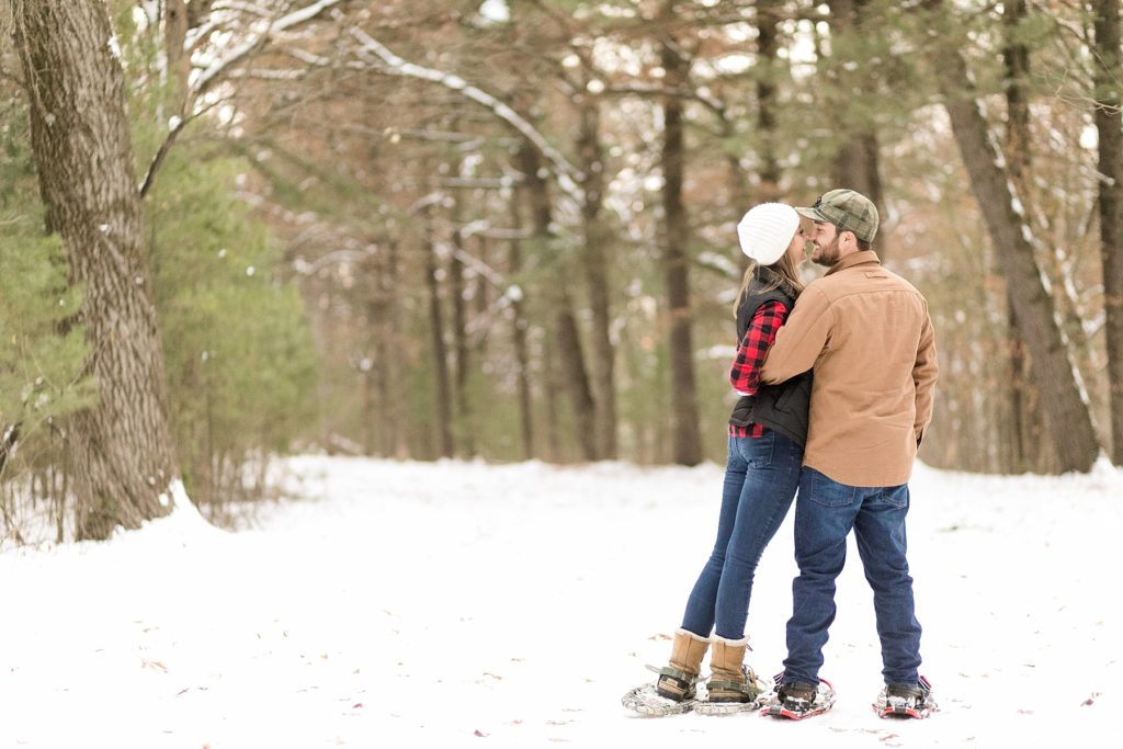 couple facing away on a snow-covered road in the woods with their snowshoes on for their snowshoe engagement photos in Wisconsin