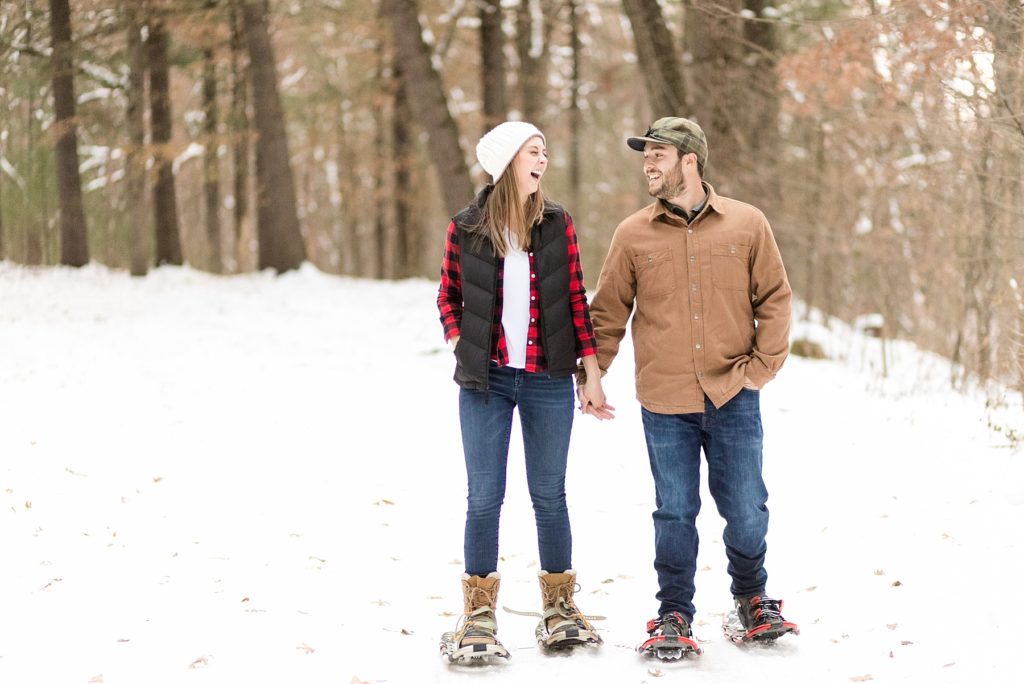 couple laughing and holding hands while snowshoeing through the woods for their snowshoe engagement session in Wisconsin