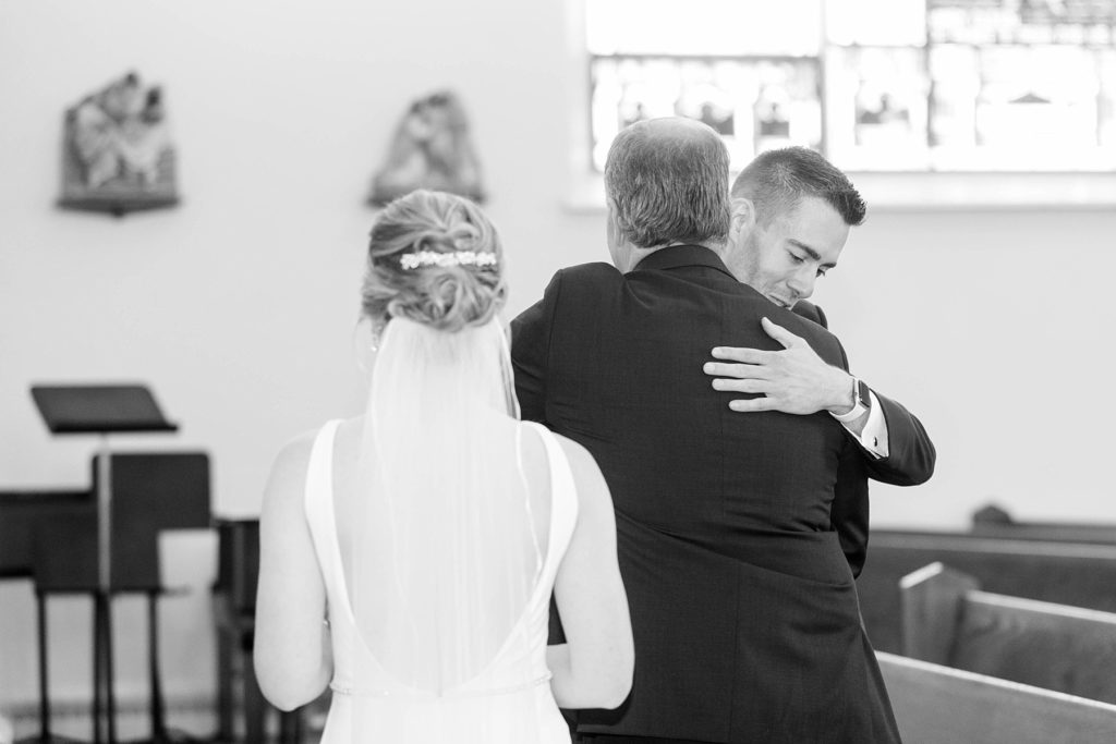groom hugging the father of the bride and bride watching in a black and white photo at St. Joseph Catholic Church in Rice Lake,. WI