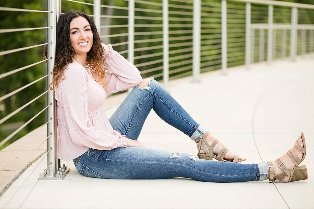 girl in a pink top and ripped jeans with tan heels leaning against a wire railing for her Eau Claire senior photos
