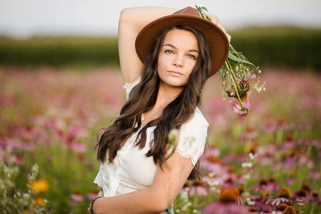 girl holding flowers with a brown hat on in a field of wildflowers for her boho senior session in Eau Claire, WI