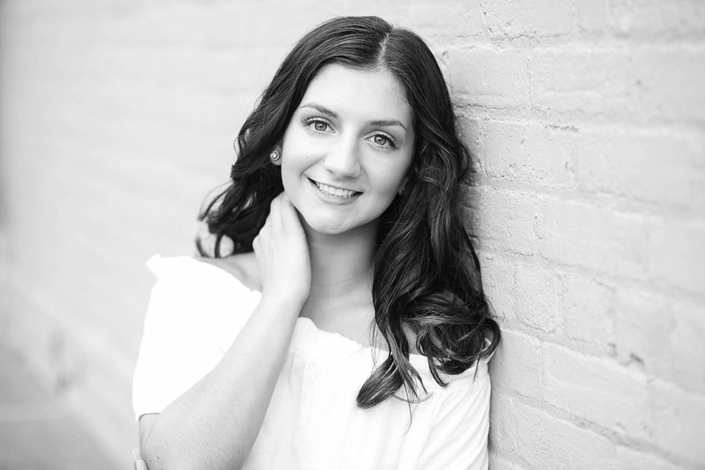 girl in a black and white photo smiling against a white brick wall for her Bloomer summer senior pictures