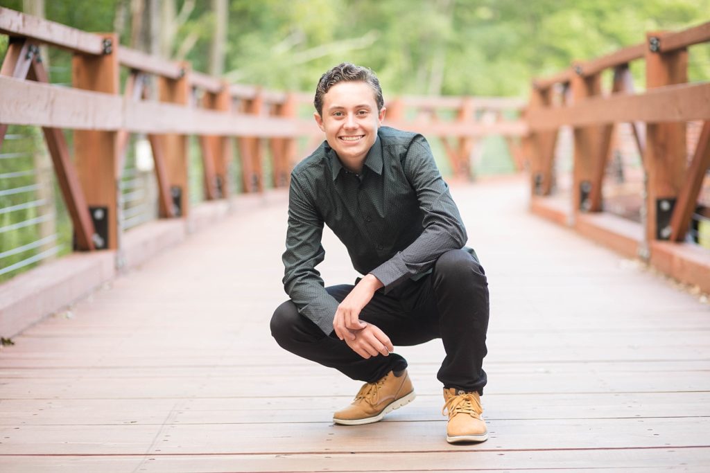 boy crouching on a bridge in a black button up shirt and black jeans at Irvine Park for his senior photos in Chippewa Falls, WI
