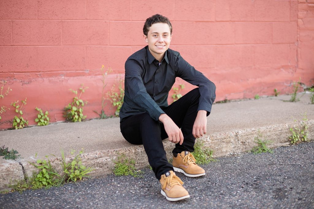 boy sitting on the curb infront of a red wall in a black outfit downtown Chippewa Falls for his senior pictures