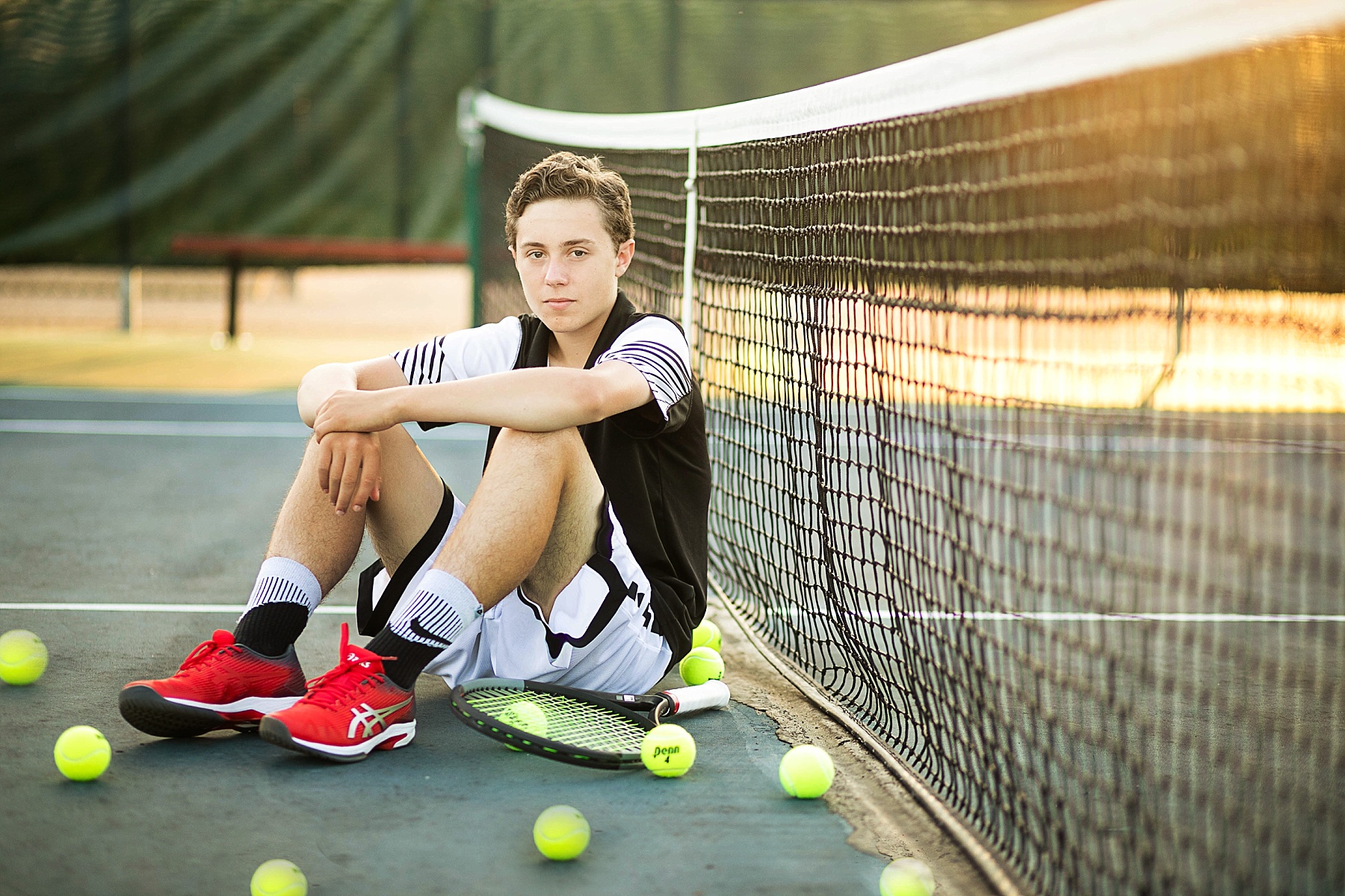 boy sitting against a tennis net at chi hi with his uniform on and tennis balls around him