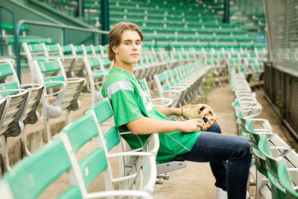 boy sitting in stands with a green Regis High School baseball jersey on in Carson Park in Eau Claire for his senior photos
