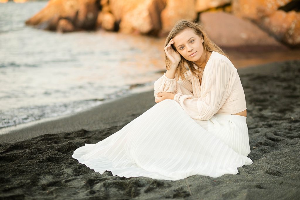 girl sits in on a black sand beach on Lake Superior with a gown on for her North Shore senior pictures