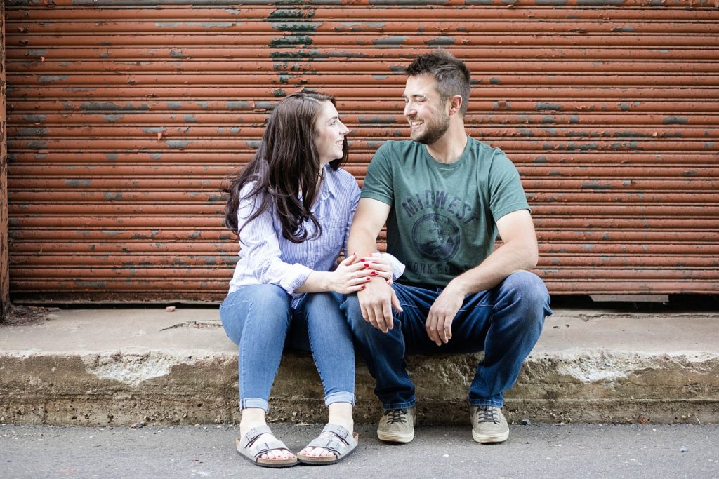 couple sitting in front of a orange garage door with peeling paint in casual spring engagement photos in Eau Claire